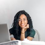Joyful businesswoman with curly hair smiling at camera while using laptop indoors.